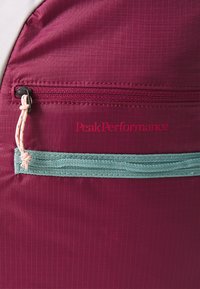 Mochila marrom com bolso frontal e zipper vermelho, cordão rosa, alça azul-petróleo e logotipo "Peak Performance" em texto vermelho.