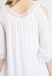 Blouse blanche avec un design épaules découpées, texture plissée et accents en dentelle aux manches et au col.