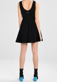 Frau in einem ärmellosen schwarzen Kleid mit schwingendem Rock und schwarzen Knöchelstiefeln mit leuchtend blauen Absätzen, steht mit dem Rücken zur Kamera.