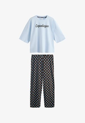 Femeie cu păr creț zâmbitoare și atingându-și capul, purtând un tricou albastru deschis "Copenhagen Frederiksberg" și pantaloni negri cu buline albe.