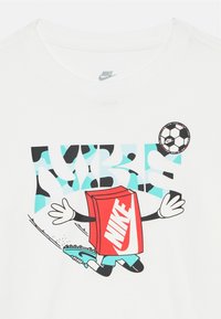 Valkoinen lyhythihainen T-paita, jossa on sarjakuvahahmo punaisella laatikkopäällä, jonka ylle on merkitty "NIKE", jalkapallo ja värikäs graafinen tausta.