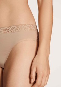Beige Slips mit einem blumenverzierten Spitzenbund, gefertigt aus glattem Stoff. Das Design zeichnet sich durch sanfte Kanten und nahtlose Verarbeitung aus.