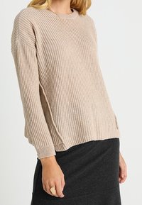 Beige värillinen ribbineulottu neulepaita, jossa pyöreä pääntie ja pitkät hihat, rento istuvuus ja sivuaukot, yhdistetty tummanharmaaseen hameeseen.