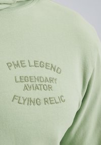 Sudadera verde claro hecha de tela suave con texto en relieve: "PME LEGEND," "LEGENDARY AVIATOR" y "FLYING RELIC." Textura suave, diseño casual.