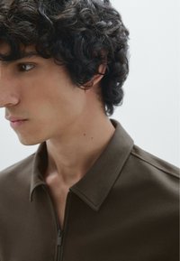 Giacca marrone con zip e colletto. Texture morbida, design minimalista. Il modello mostra un profilo laterale su uno sfondo neutro.