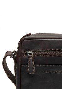 Borsa in pelle marrone con texture liscia, tasca con cerniera e tracolla regolabile. Presenta un branding discreto e dettagli di cucitura.