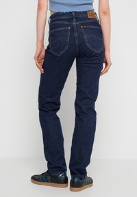 Vue de dos d'une personne portant un jean droit bleu foncé, un haut court bleu clair et des baskets noires avec des rayures bleues et des semelles en gomme.