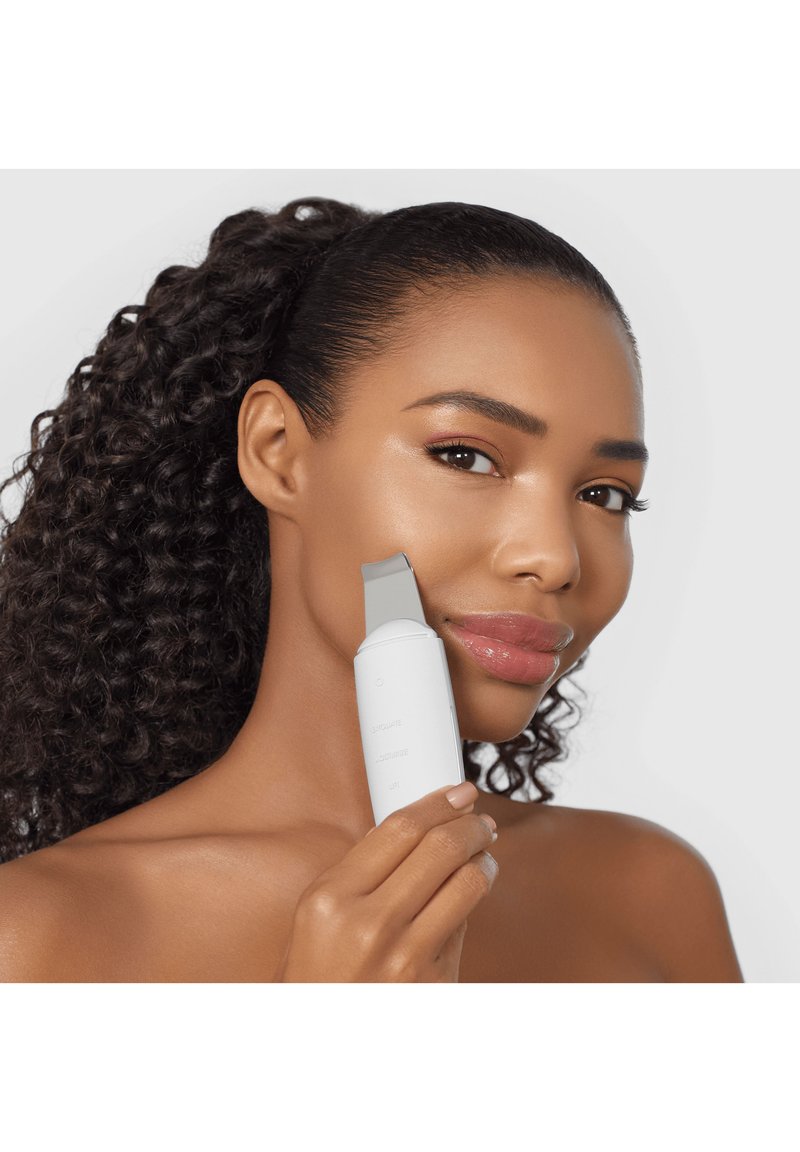 Woman with curly hair using a white facial cleansing device on her cheek, demonstrating skincare routine on a neutral background.