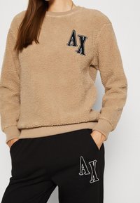 Felpa beige pelosa con scollo tondo, polsini e orlo a coste. Presenta il logo "AX" ricamato in nero sul lato sinistro del petto. Pantaloni neri con logo abbinato.