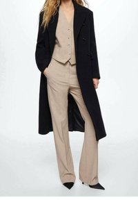 Costume beige sur mesure avec un gilet ajusté et un pantalon à jambes larges, superposé sous un long manteau noir, doté de boutons double boutonnage et d'une silhouette épurée.