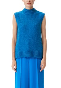 Ärmelloses blaues Strickoberteil mit hohem Kragen, das über eine strukturierte Oberfläche und einen gerippten Saum verfügt. Getragen über einem passenden blauen, fließenden Rock.