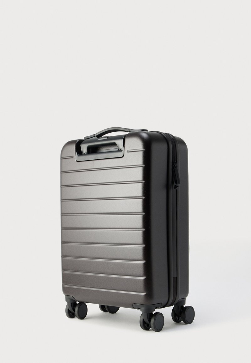 Hard-shell carry-on suitcase in metallic dark gray, featuring horizontal ridges, top handle, and four rotating wheels for mobility.