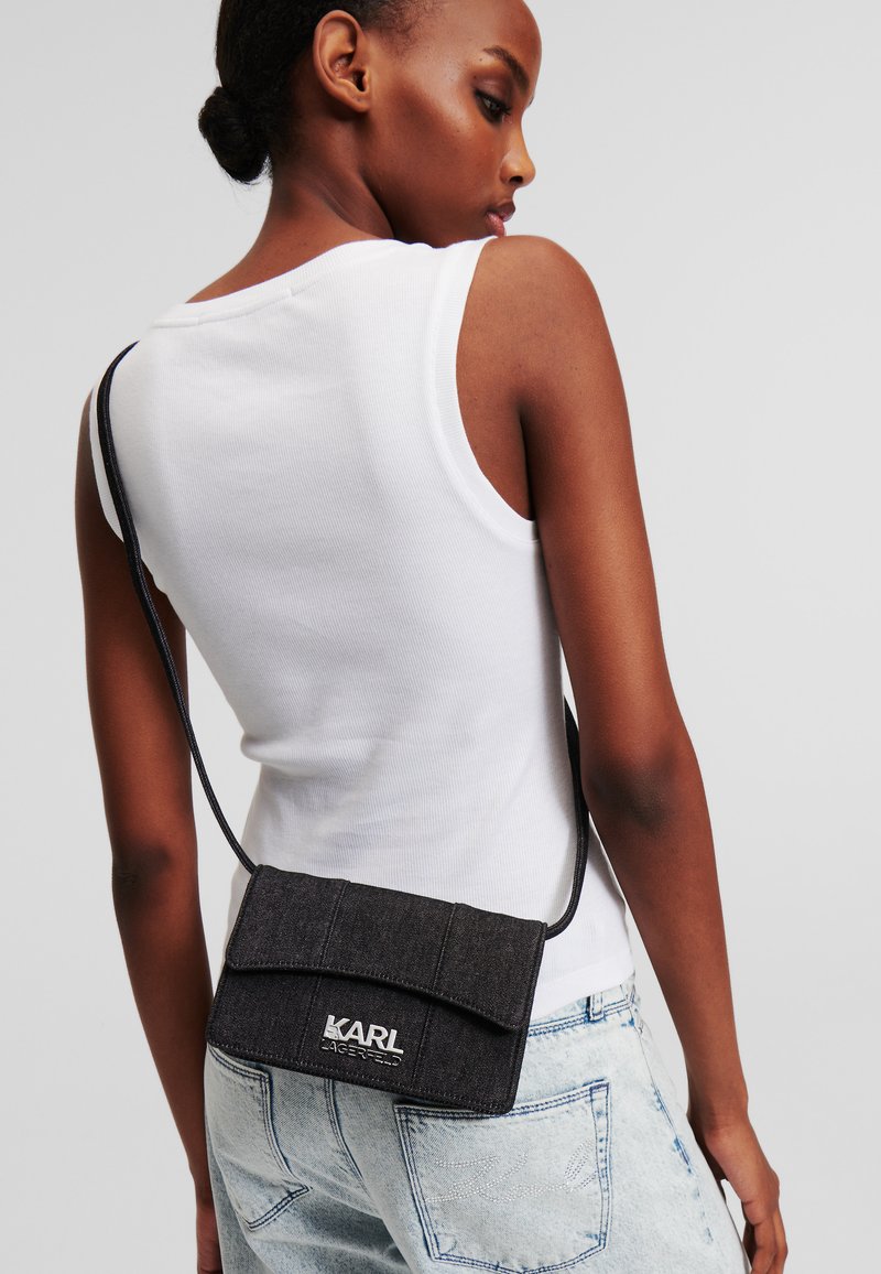 Sac en denim noir porté en bandoulière avec un rabat, arborant un logo argenté, associé à un jean bleu clair et un débardeur blanc.