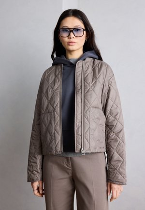 Mlada ženska, oblečena v sivo odejano jakno preko temno sive kapucarice in bež hlače, z velikimi toniranimi očali, stoji pred enostavnim sivim ozadjem.