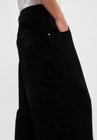 Fekete kordbársony nadrág függőleges bordázott textúrával, zsebeknél koponya és keresztcsont részlettel. Oldalzseb látható.