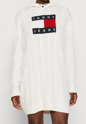 Biała sukienka sweterkowa z kapturem, z splotem warkoczowym, z logo Tommy Jeans w kolorach czerwonym, białym i niebieskim na piersi, noszona przez osobę o ciemnym odcieniu skóry.