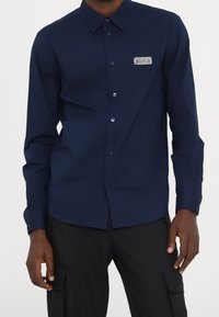 Maglietta a maniche lunghe blu navy realizzata in tessuto morbido, con chiusura frontale a bottoni, colletto a punta e una patch con logo sul lato sinistro.