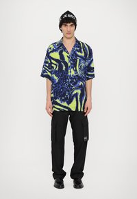 Trumpas rankovėmis marškiniai su mėlyna ir žalios citrinos spalvos abstrakčia raštine; dėvimi su juodomis kargo kelnėmis ir juodu beanie, ant kurio užrašyta balta tekstas.