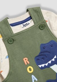 Grüne Baumwolloveralls mit einem Dinosaurierdesign und buntem "ROAR"-Stickerei, mit Knopfverschlüssen an den Trägern und einem hellbeigen Innenhemd.