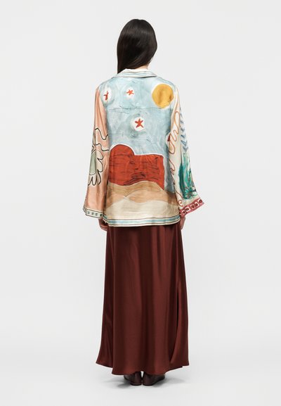 Γυναίκα που φαίνεται από πίσω, φοράει μπλούζα μακρυμάνικη με πολύχρωμα αφηρημένα τοπία και μοτίβα αστερία, και μια μακριά φούστα μαξι από καφέ σατέν.
