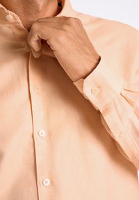 Helles Pfirsichfarbenes Langarmhemd mit Button-Down-Kragen, Ein-Knopf-Manschetten und sichtbaren Vorderknöpfen. Der Stoff wirkt weich und strukturiert.