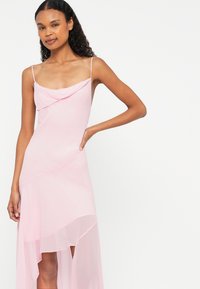 Ružové šaty s drapovaným výstrihom, tenkými ramienkami a asymetrickým lemom. Vyrobené z ľahkého materiálu s priesvitnou spodnou časťou.