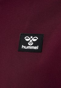 Burgunderfarvet stof med en sort rektangulær lap med et hvidt logo og teksten "hummel" prægede i midten. Glat tekstur, minimalistisk design.
