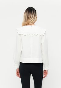 Blusa bianca con colletto arricciato e maniche lunghe. Il tessuto appare liscio e leggero, con una cerniera sul retro.