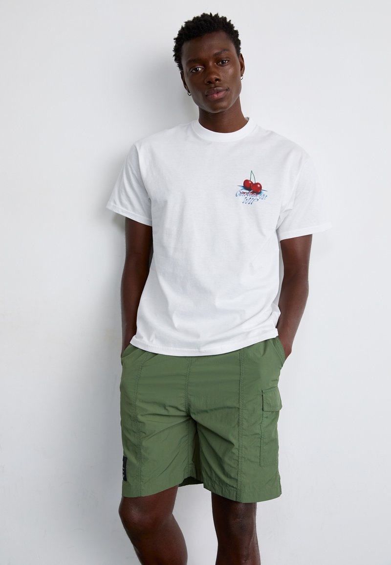 Baltas medvilninis marškinėlis su vyšnios piešiniu ant krūtinės, derinamas su žaliomis krovinių šortais iš lengvos medžiagos.