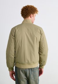Khaki bomber bunda s hladkým povrchom, s elastickými manžetami a pásom. Zobrazuje klasický siluetu a minimalistický dizajn, vhodný na casual nosenie.