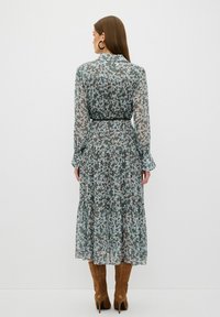 Kwiatowa midi sukienka z długimi rękawami, przezroczystym materiałem, zielono-niebieskim wzorem, z wcięciem w pasie, warstwową spódnicą, zestawiona z brązowymi botkami na obcasie.