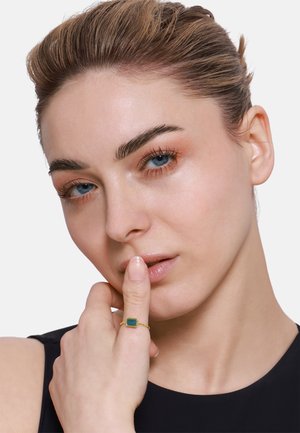 Goldring mit einem rechteckigen grünen Stein, an der rechten Hand getragen. Nahaufnahme eines Models mit heller Haut und zurückgegeltem Haar, das ein schwarzes Oberteil trägt.