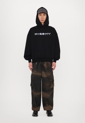 Svart oversized hettegenser med "MISBHV" tekst; sammen med løse cargo-bukser i brunt og svart med lommer; båret med en grå caps og svarte sko.