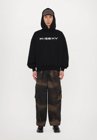 Zwarte oversized hoodie met "MISBHV" tekst; gecombineerd met losse cargobroek in bruin en zwart met zakken; gedragen met een grijze pet en zwarte schoenen.