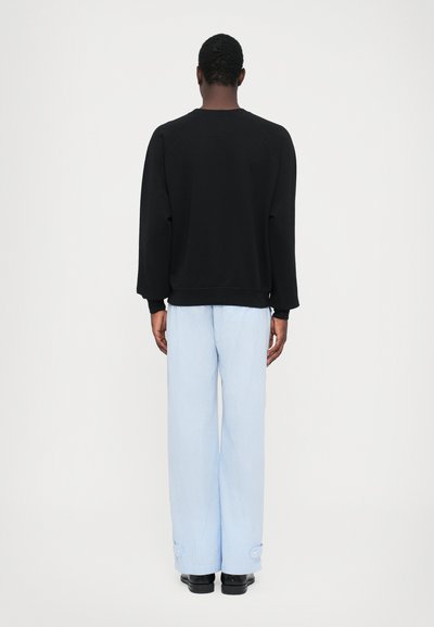 Schwarzer Sweatshirt mit lockerer Passform und gerippten Bündchen; hellblaue gestreifte weit geschnittene Hose mit Knopfdetail an den Bündchen; schwarze Schuhe.