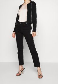 Femme portant une veste noire zippée, un haut en satin rose clair, un jean noir, des sandales à talons hauts imprimé serpent, et tenant une pochette blanche à perles.