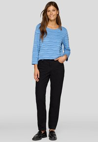 Helles blaues Oberteil mit strukturiertem Wellenmuster, kombiniert mit schwarzen, geraden Hosen und schwarzen Loafers. Einfaches Design, lässige Passform.