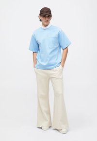 Noor mees, kes kannab sinist üle mõõdu rõivast, kreemjat laia püksipaari, valgeid jalatseid ja tumedat pruuni mütsi, seistes käed taskus.