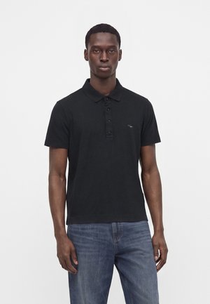 Giovane uomo con indosso una polo nera a maniche corte e jeans blu, in piedi su uno sfondo bianco semplice.