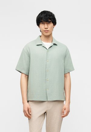 Jeune homme portant une chemise boutonnée à manches courtes vert clair sur un t-shirt blanc et un pantalon beige, debout devant un fond uni.