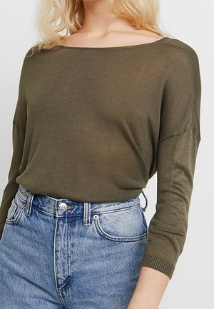 Olivgrüner Strickpullover mit weitem Ausschnitt und Dreiviertelärmeln, kombiniert mit hoch geschnittenen hellblauen Jeans.