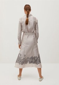 Donna con capelli intrecciati, che indossa un vestito bianco a pois con stampa a paisley sul bordo, in posizione di spalle, abbinato a scarpe slip-on bianche.