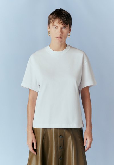T-shirt bianco in cotone con scollo rotondo e maniche corte, abbinato a una gonna in pelle marrone con dettagli in bottoni. Texture liscia, design casual.