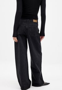 Črne široke hlače iz jeansa z zdblebeno teksturo, petimi žepi in blagovno znamko v usnjenem bež pečatu na pasu. Okrašene z majhnimi žebljički.