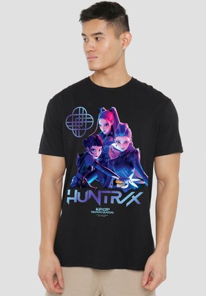 Mann trägt ein schwarzes T-Shirt mit einem farbenfrohen Motiv von drei animierten weiblichen Charakteren und dem Text "HUNTR/X K-POP DEMON HUNTERS."