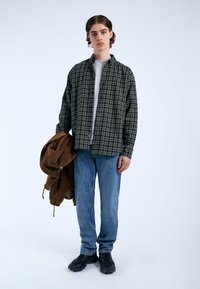 Camisa a cuadros negra y crema, camiseta gris, jeans de mezclilla azul claros y zapatillas oscuras. Sosteniendo una chaqueta de pana marrón. Diseño simple y casual.