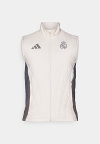 Chaleco sin mangas blanco y azul marino con alto cuello, ligero acolchado, cremallera frontal, logo de Adidas y emblema del Real Madrid en el pecho.