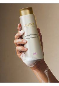 Witte fles AHAVA Mineral Shampoo met gouden dop, gehouden in een zeepproep. Het etiket bevat zwarte tekst en het woord "HAIR" in het roze.