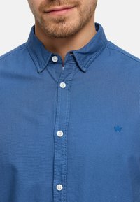 Partasuuinen mies sininen nappipaita päällä, jossa on pieni kirjailtu logo rinnassa, kaulus osittain auki.