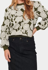 Olivgrüner gestickter Pullover mit weißen Blumenmuster, strukturiertem Stoff, gerippten Bündchen und Saum. Kombiniert mit einem dunklen Jeansrock.